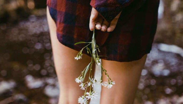Woman in plaid dress holding small white flowers behind her back outdoors related to causes of incontinence awareness