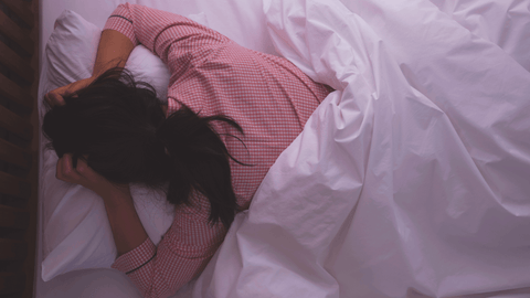 Woman in pink pajamas lying on bed with white sheets, struggling to sleep, representing hormonal sleep disruptions and restful sleep challenges.